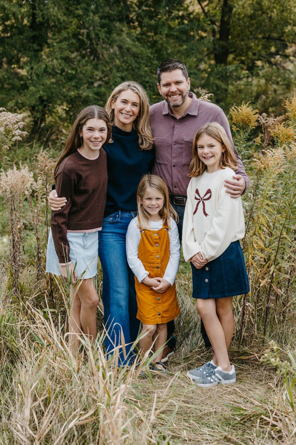 A family of five, including two adults and three young girls, stands together outdoors in a grassy area, smiling at the camera.