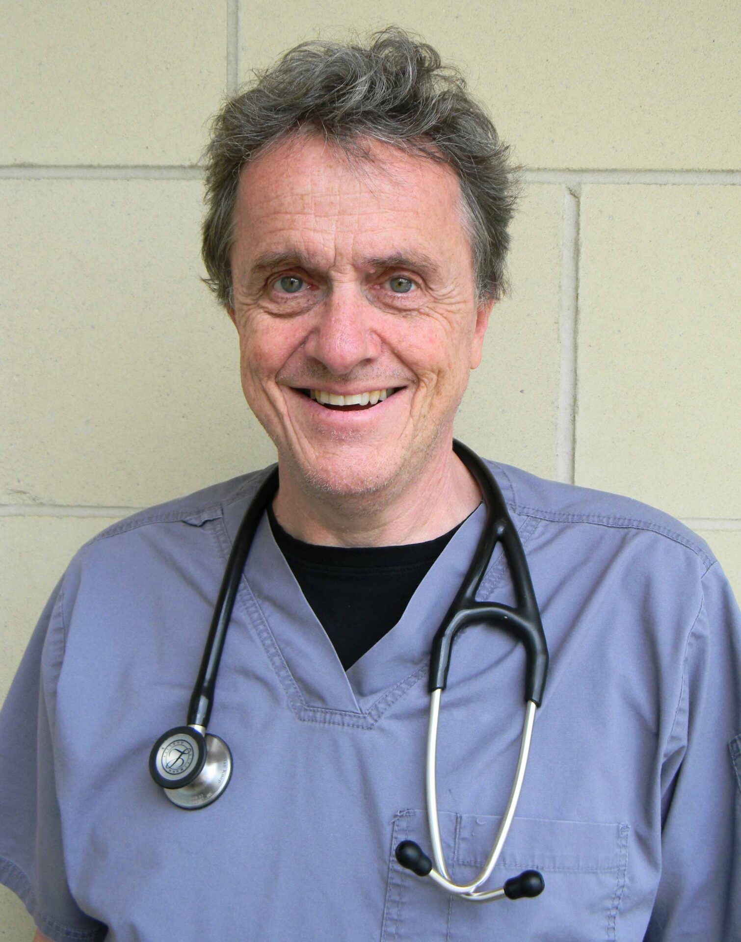A person in grey scrubs with a stethoscope around their neck stands in front of a beige tiled wall, smiling at the camera.