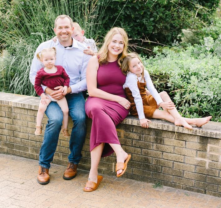 A family of five sits on a brick ledge outdoors; two adults and three young children pose and smile at the camera, surrounded by greenery.