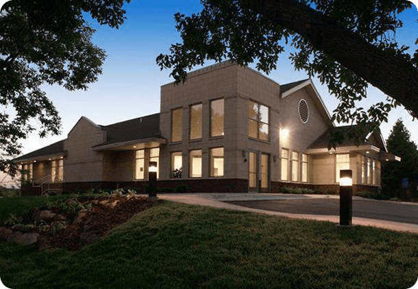 A modern one-story building with large windows and exterior lights, surrounded by grass, trees, and a driveway at dusk.