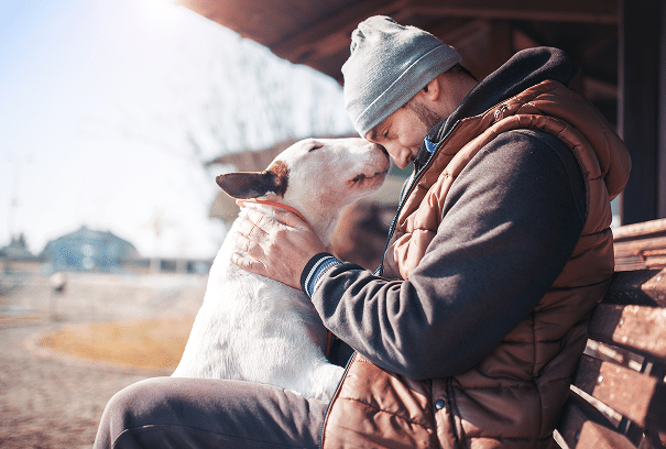 A man in a beanie and vest sits on a bench, touching foreheads with a white bull terrier dog in an outdoor setting.