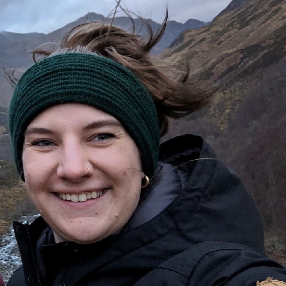 A person wearing a dark headband and jacket smiles outdoors with mountains in the background on a cloudy day.