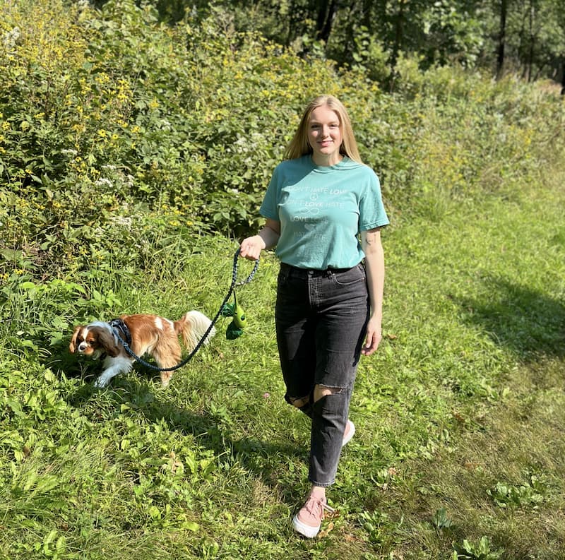 A woman walks a brown and white dog on a grassy path surrounded by green foliage on a sunny day.