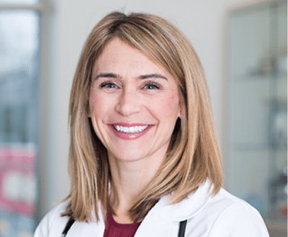 A woman with straight, shoulder-length blonde hair, wearing a white medical coat and smiling, stands in a bright, indoor setting.