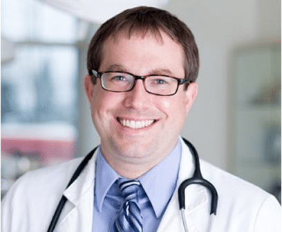 A male doctor wearing glasses, a white coat, a blue shirt with a striped tie, and a stethoscope, smiling in a medical office.