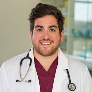 A person wearing a white lab coat and maroon scrubs with a stethoscope around their neck, standing indoors and smiling at the camera.