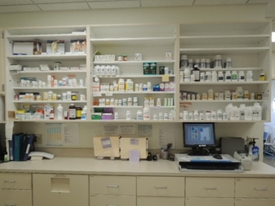 Shelves filled with various prescription medication bottles above a counter with folders, a computer, and a printer in a pharmacy workspace.