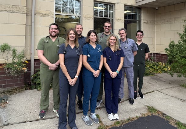 Eight healthcare professionals wearing scrubs and stethoscopes stand outside a building, facing the camera and smiling.