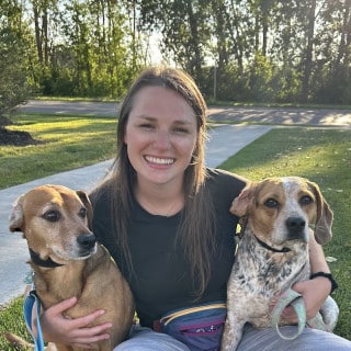 A woman sits on grass smiling, holding two dogs on leashes, with trees and a sidewalk in the background.