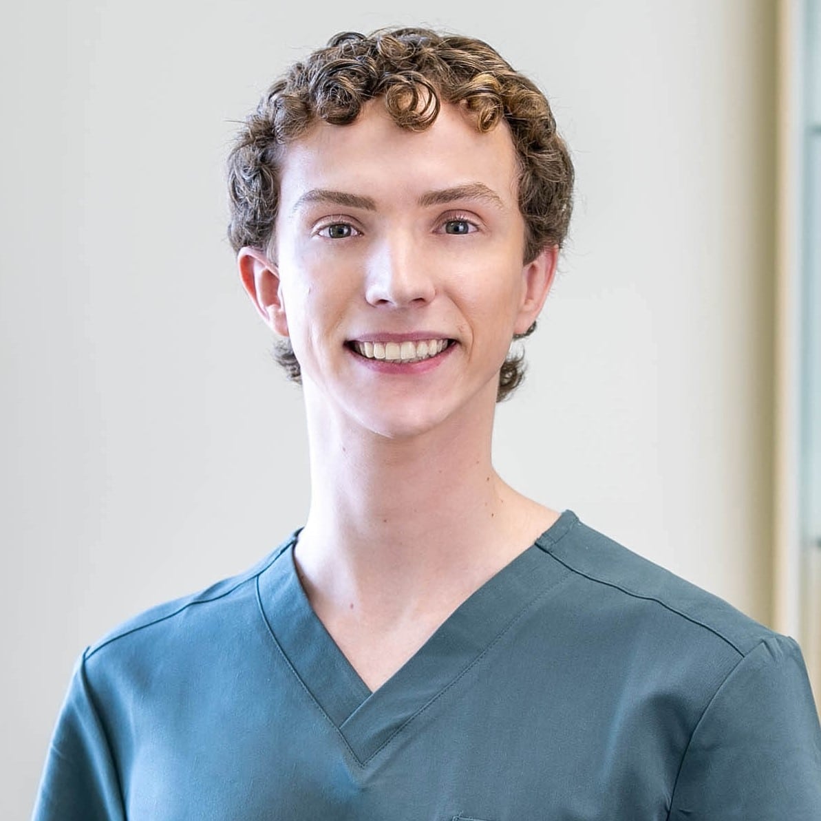 A person with short curly hair, wearing teal medical scrubs, stands indoors and smiles at the camera.