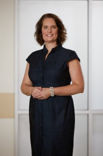 Woman with short brown hair wearing a dark, short-sleeved dress stands indoors, smiling, with hands clasped in front of her.