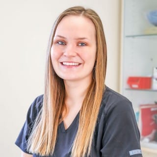 A woman with long straight hair wearing medical scrubs smiles while seated in a clinical setting with shelves in the background.