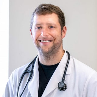 A man wearing a white lab coat and a stethoscope around his neck stands smiling in front of a plain background.