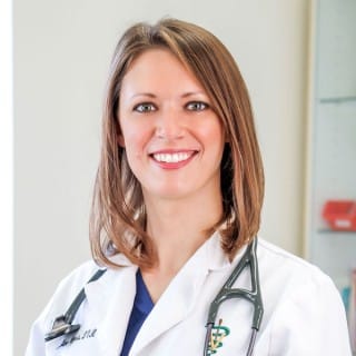 A woman in a white medical coat with a stethoscope around her neck is smiling at the camera in a clinical setting.