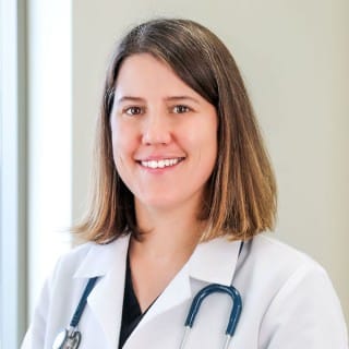 A woman in a white medical coat with a stethoscope around her neck stands indoors, smiling at the camera.