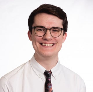 A person with short dark hair, wearing glasses, a white shirt, and a floral tie smiles in front of a plain white background.