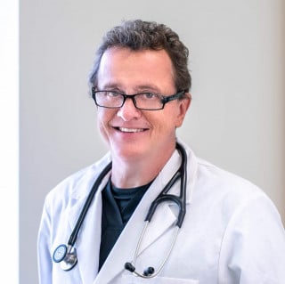 A middle-aged male doctor with glasses and a stethoscope around his neck, wearing a white lab coat, smiles at the camera.