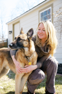 A woman kneels outside next to a large German Shepherd dog in front of a light-colored house on a sunny day.