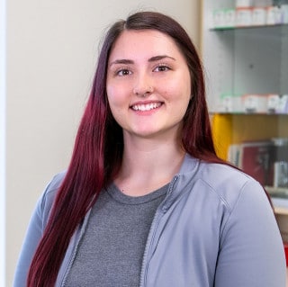 A young woman with long dark hair and a nose ring smiles, standing indoors near shelves containing various items.
