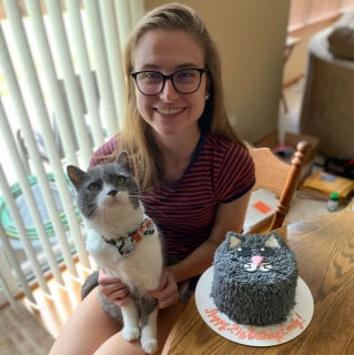 A woman wearing glasses holds a gray and white cat beside a cake decorated to look like the cat's face, on a wooden table indoors.