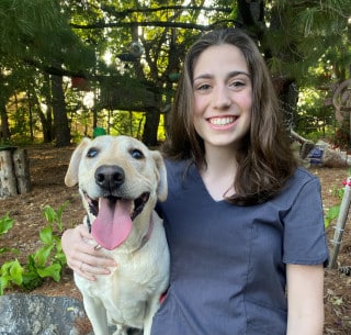 A young woman in gray scrubs sits outdoors next to a yellow Labrador retriever with its mouth open and tongue out, both looking at the camera. Trees and plants are in the background.