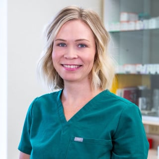 A woman in green medical scrubs stands in front of shelves with medical supplies, smiling at the camera.