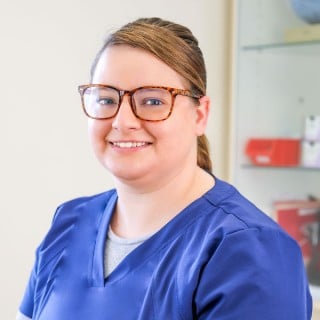 A woman wearing glasses and blue scrubs is smiling at the camera in a well-lit indoor setting with medical supplies in the background.