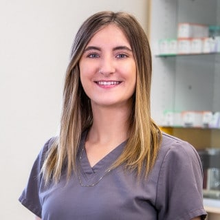 A woman wearing gray scrubs stands in front of pharmacy shelves, smiling at the camera.