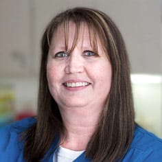 A woman with straight brown hair, wearing a blue top, smiles at the camera in an indoor setting with a light-colored background.