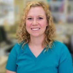 A woman with curly blonde hair wearing teal scrubs smiles at the camera in an indoor setting with a blurred background.
