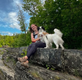 A woman sits on rocky terrain outdoors with two small dogs in front of a backdrop of green trees and a partly cloudy sky.