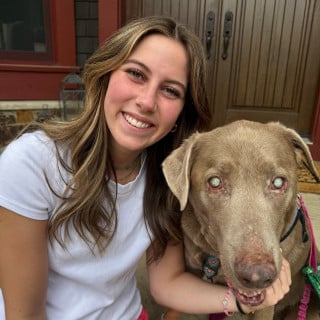 A young woman in a white t-shirt smiles next to a brown dog with cloudy eyes, sitting outside in front of a wooden door.