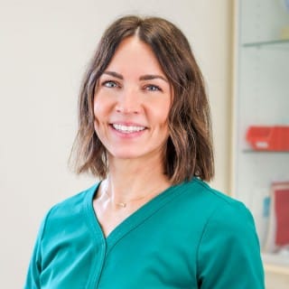 A woman with shoulder-length brown hair, wearing a teal medical scrub top, stands indoors and smiles at the camera.