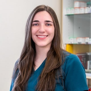 A woman with long brown hair in teal scrubs smiles at the camera, standing in front of shelves with medical supplies.