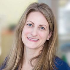 A woman with long, straight, light brown hair and blue eyes smiles at the camera. She is wearing a dark top, and the background is softly blurred.