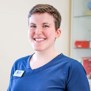 A person with short hair wearing blue medical scrubs and a name badge stands indoors, smiling at the camera.