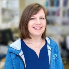 A woman with shoulder-length brown hair wearing a blue top and blue jacket smiles at the camera in an indoor setting.
